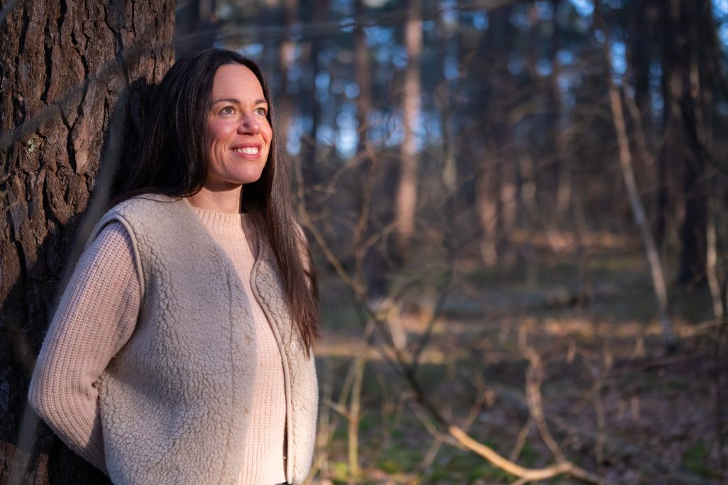 Frau mit langen Haaren lehnt an einem Baum im Wald, lächelt im Sonnenlicht.